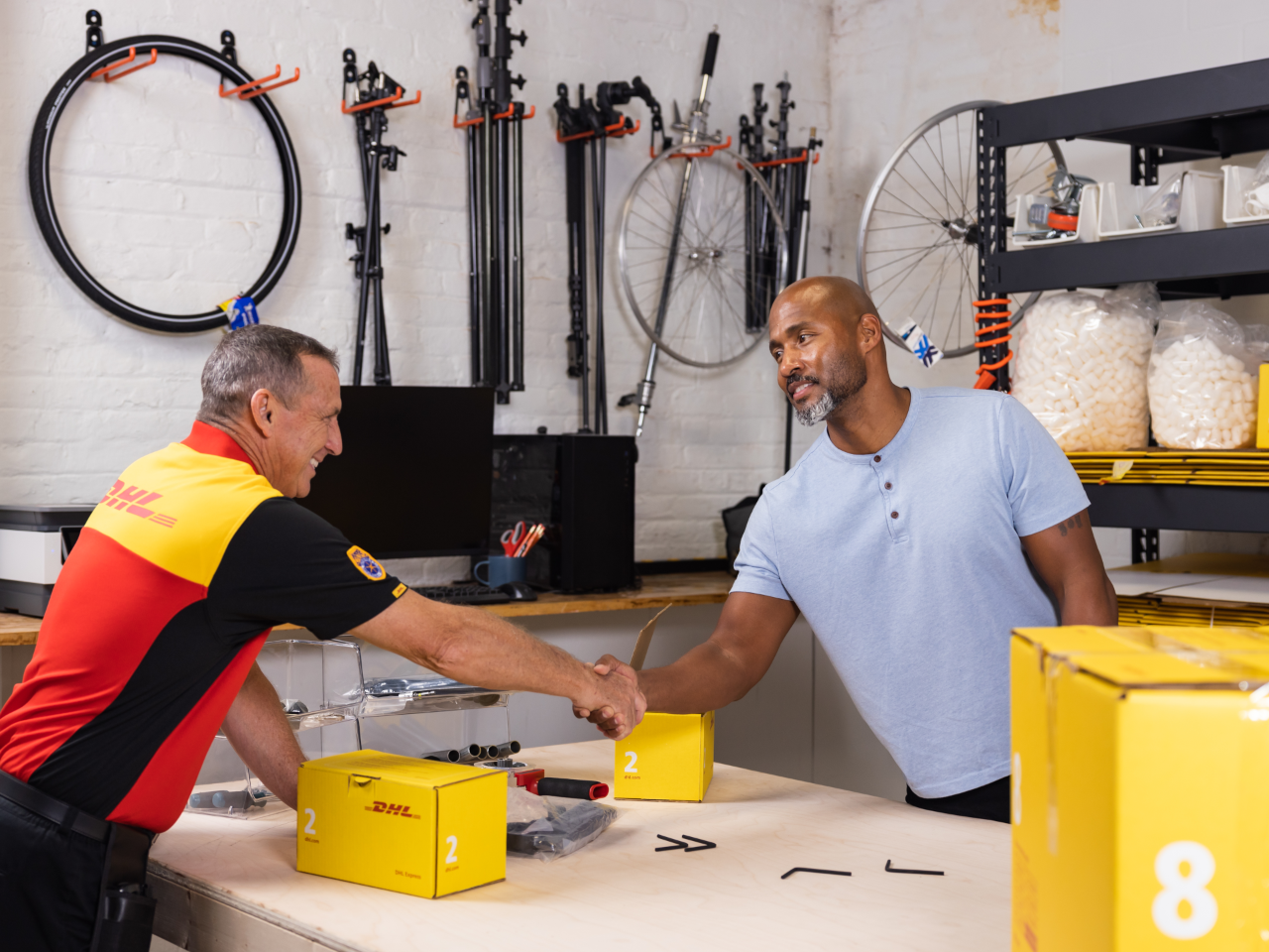 Two men are shaking hands across a workbench in a warehouse. One man is wearing a DHL shirt while holding a package, highlighting their affordable small business shipping solutions, while the other is wearing a blue shirt. Various equipment and bicycle parts are visible in the background.