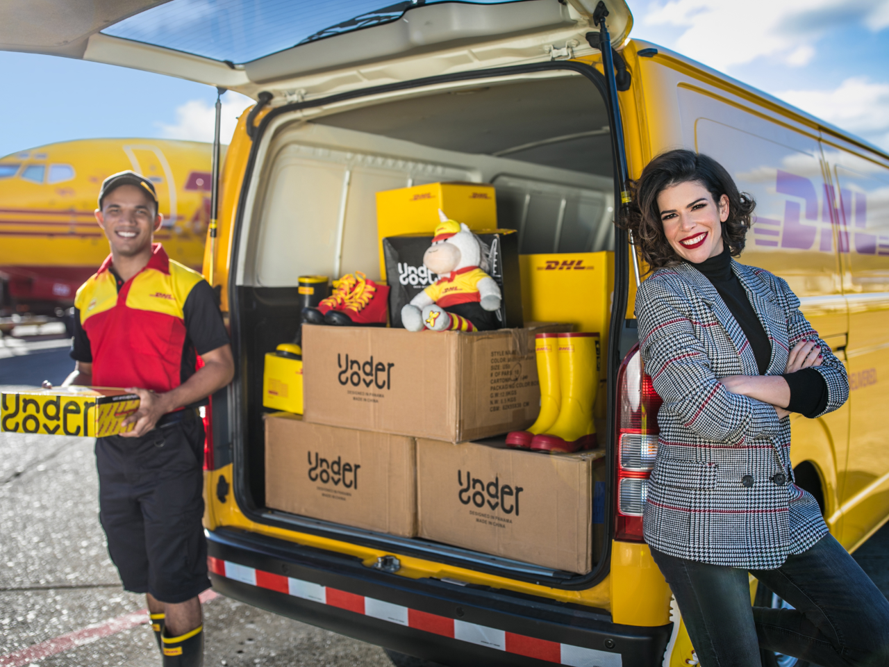 A smiling DHL worker stands next to an open DHL van filled with packages, while a woman in a gray blazer leans against the van. In the background, a DHL aircraft symbolizes their commitment to affordable small business shipping solutions.