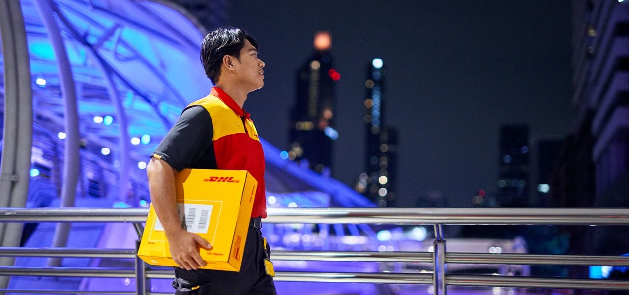 A male DHL employee walks across a bridge in a city at night with a parcel under his arm.