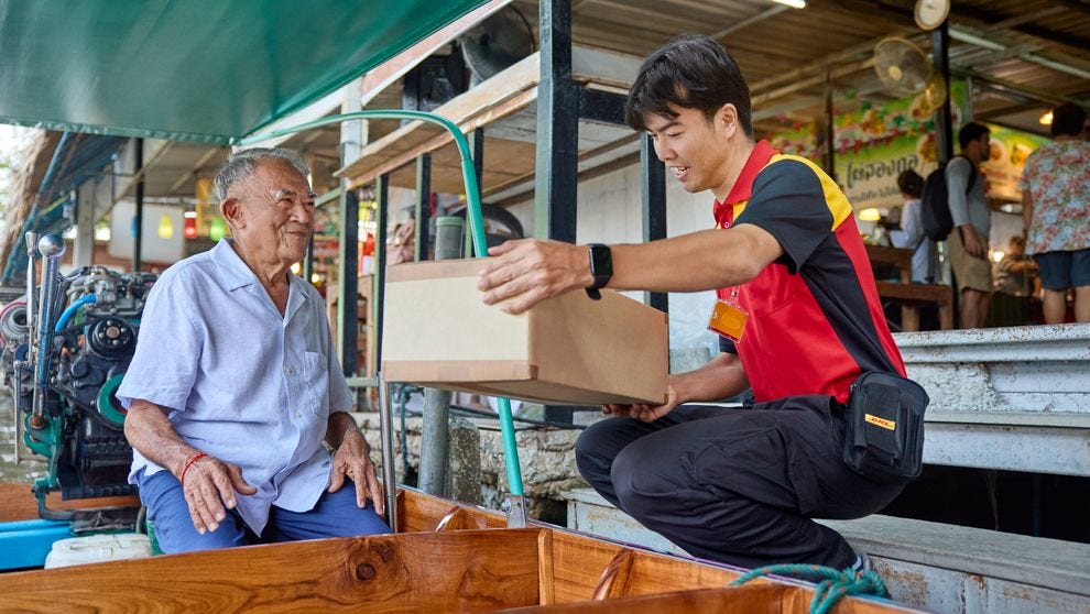 A male DHL employee hands over a parcel to an elderly man on his longtail boat. In the background, a traditional market with numerous visitors is visible.