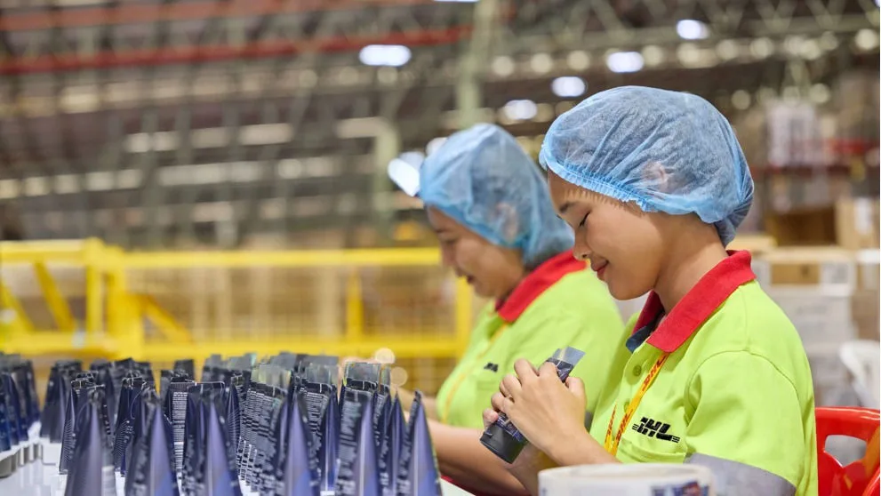 两名 DHL 仓库女工佩戴防尘帽，在仓库工位查验护肤类管状货品