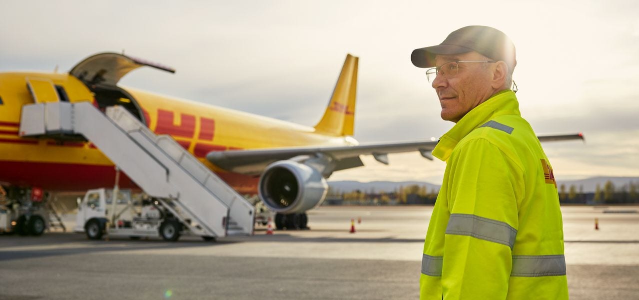 A male DHL employee standing on the airfield in front of a DHL plane