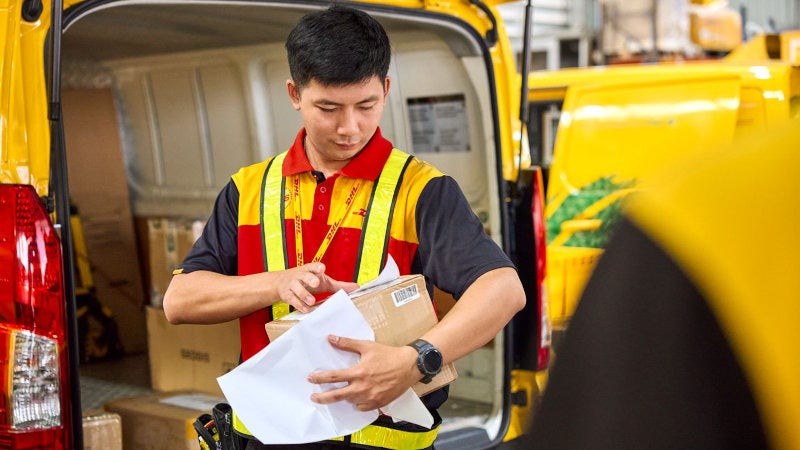 dhl employee standing in front of the open trunk of an electric dhl van and attaches a shipping label to a parcel
