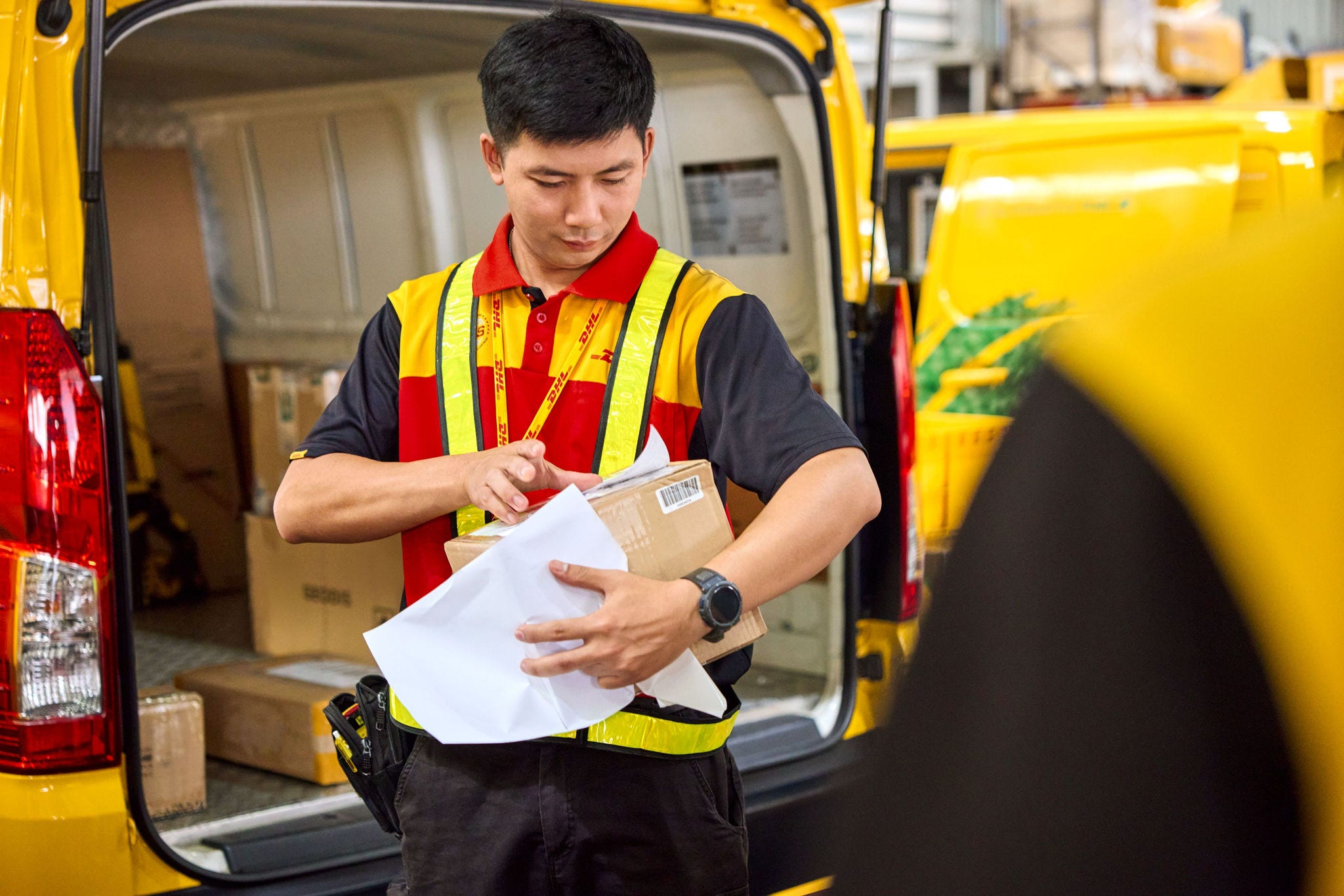 dhl employee standing in front of the open trunk of an electric dhl van and attaching a shipping label to a parcel