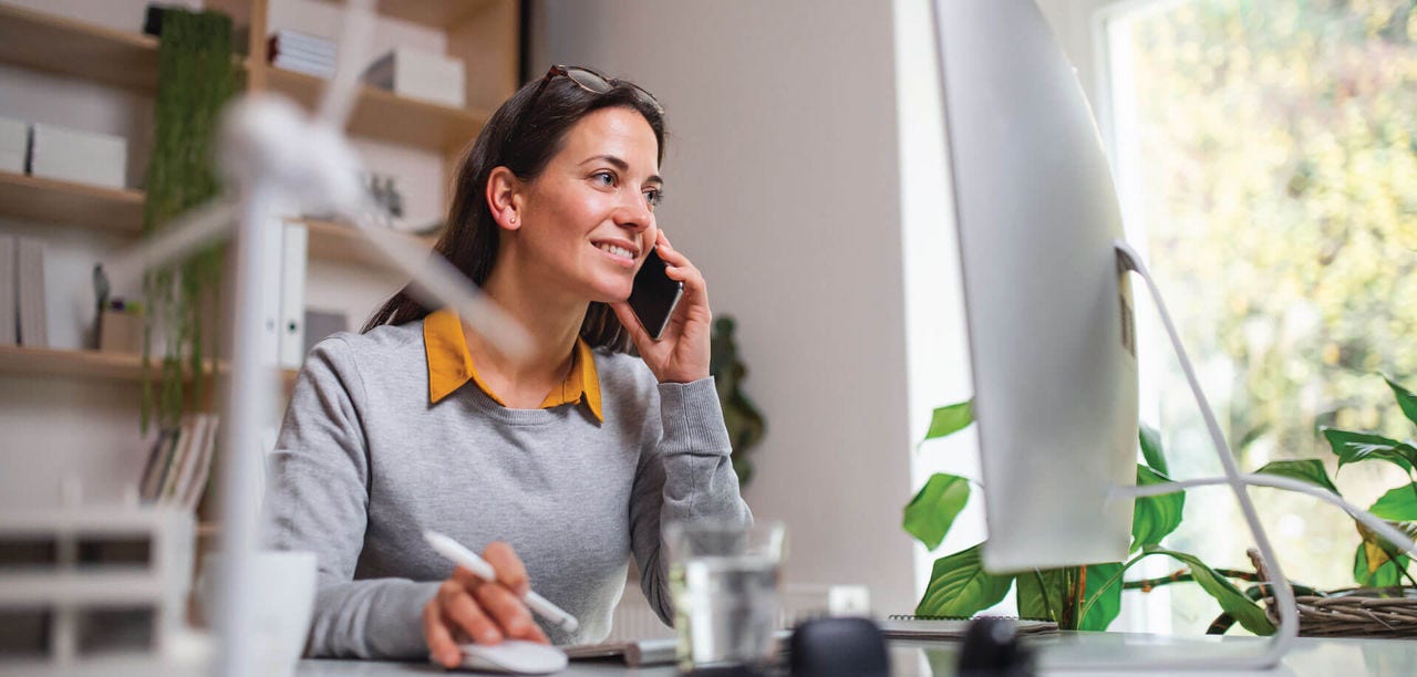 woman on her cell phone while on her computer at a desk 