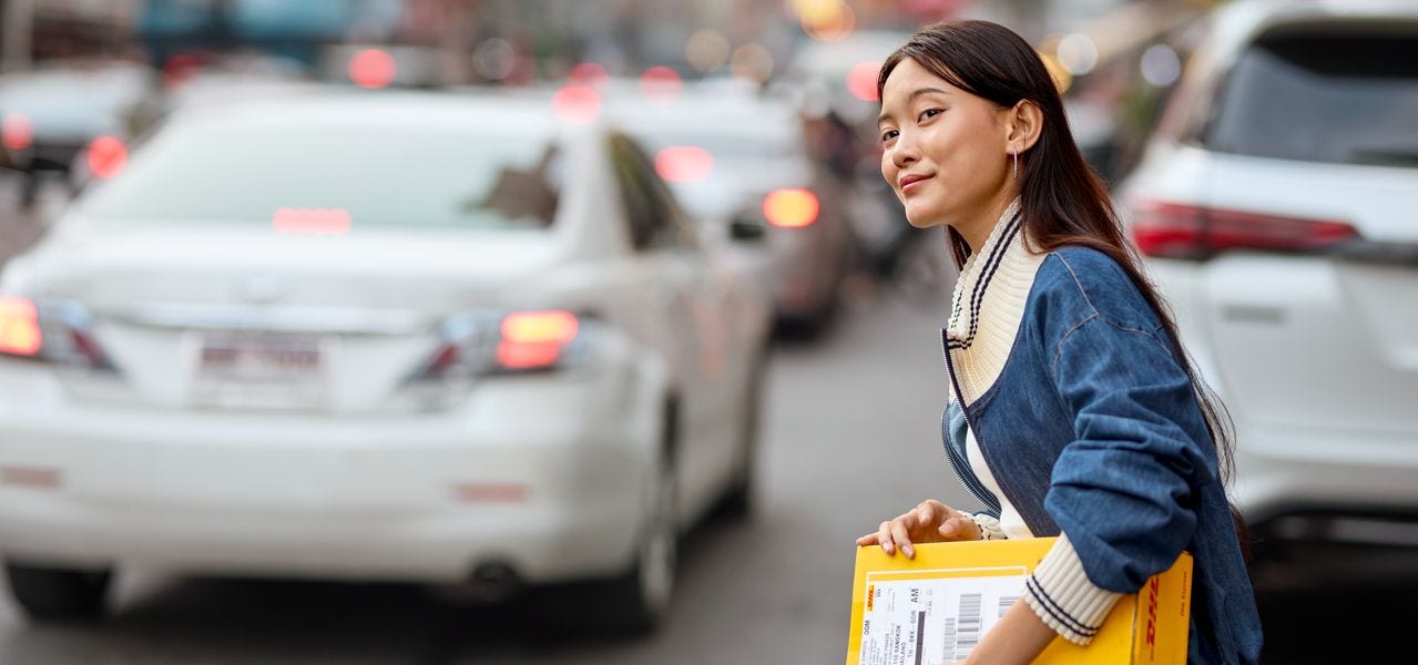 A young woman holding a DHL express parcel as she crosses a busy street in Chinatown. She glances to the side to observe the traffic.