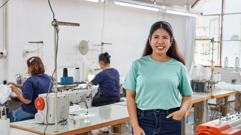 A female staff member from a supplier facility smiles at the camera while two seamstresses work on sewing machines behind her.