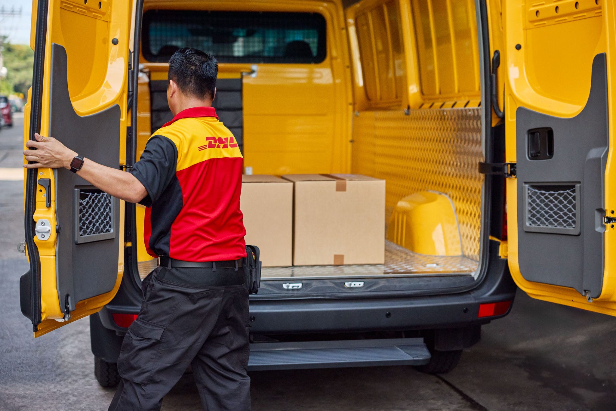 A male DHL employee loads brown parcels into the cargo area of his electric DHL van.