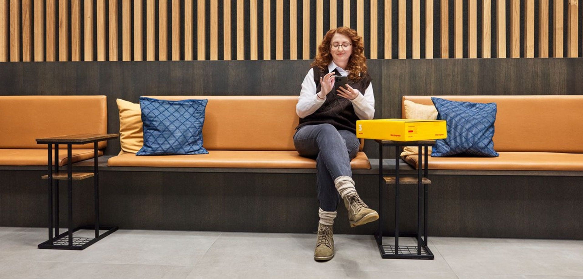 A female customer is checking her smartphone in a business building. In front of her, there is a yellow DHL express parcel at a table.