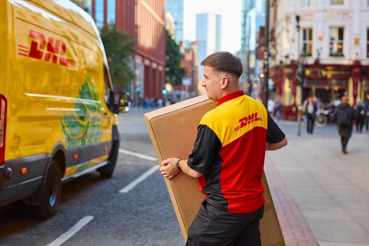 male dhl employee walking towards his electric dhl van with a brown parcel in his hand