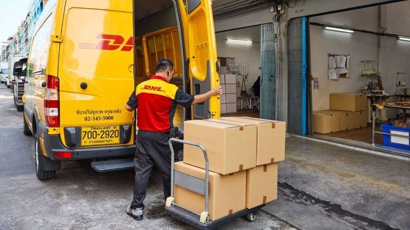 a DHL courier loading parcels from a trolley into a yellow DHL delivery van outside a warehouse.