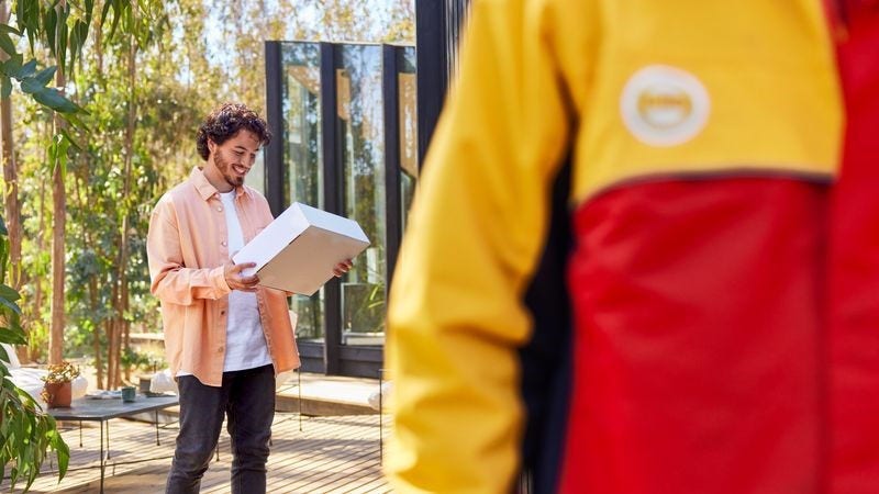 A male customer holds a parcel that has just been delivered by a DHL employee, who is walking away in the foreground.