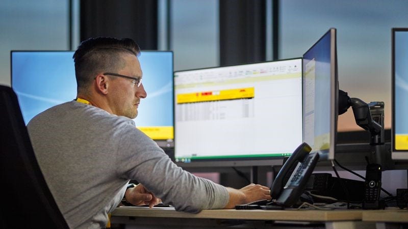 a dhl employee sits at the desk, looking at three computer screens