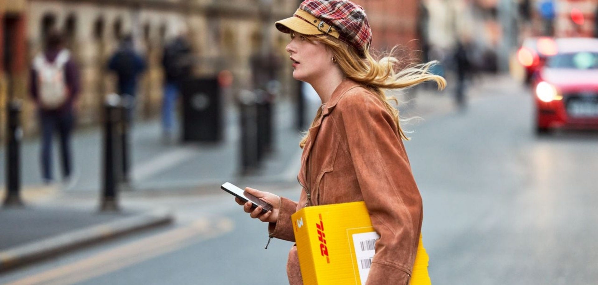 A female customer with an express parcel crosses the street. In her hand, she carries a smartphone.