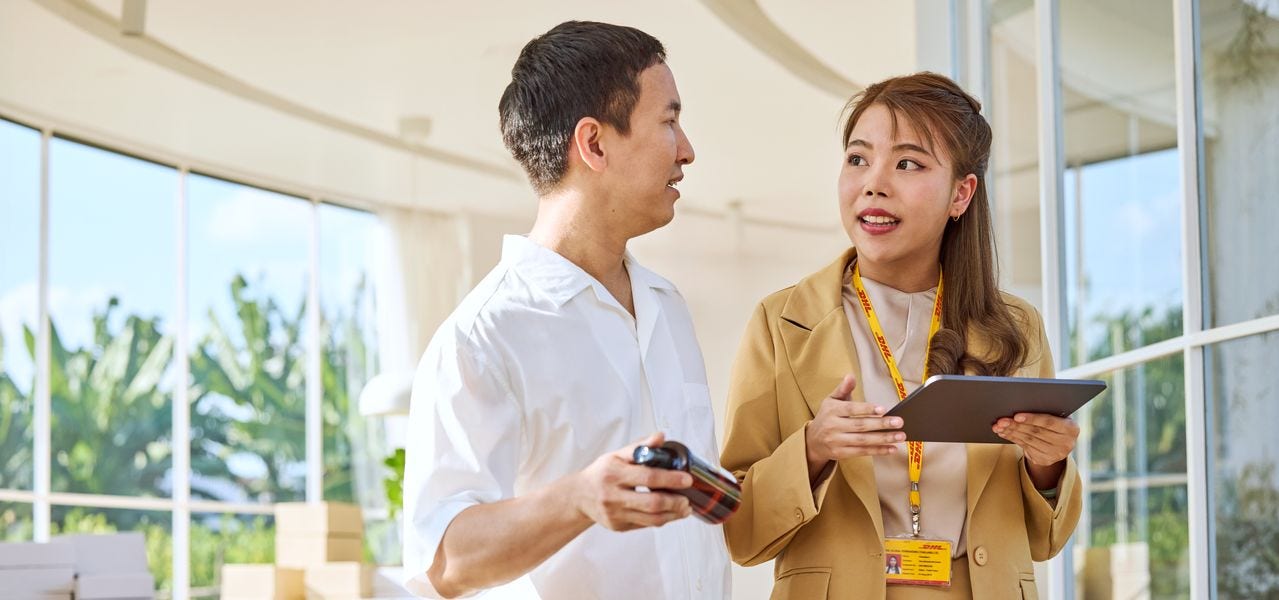 A male shop owner is showing around a female DHL expert in his store to give her an idea of the products he wants to ship with DHL