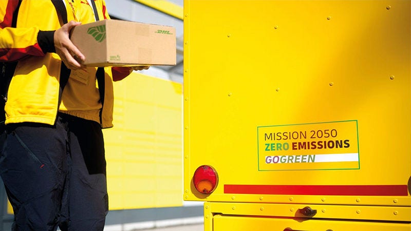 A DHL delivery person holding a package next to a DHL vehicle with a 'Mission 2050 Zero Emissions GoGreen' decal.