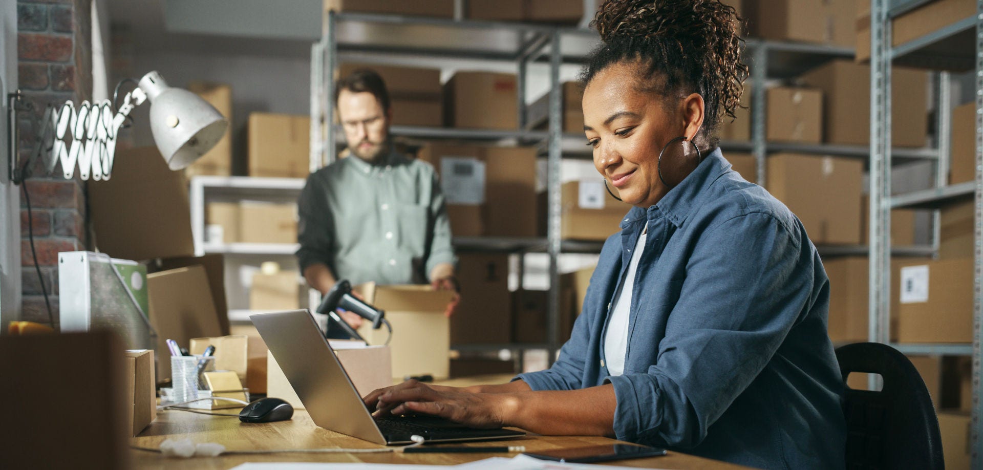 Woman sitting at a desk within a warehouse full of boxes, typing on her computer