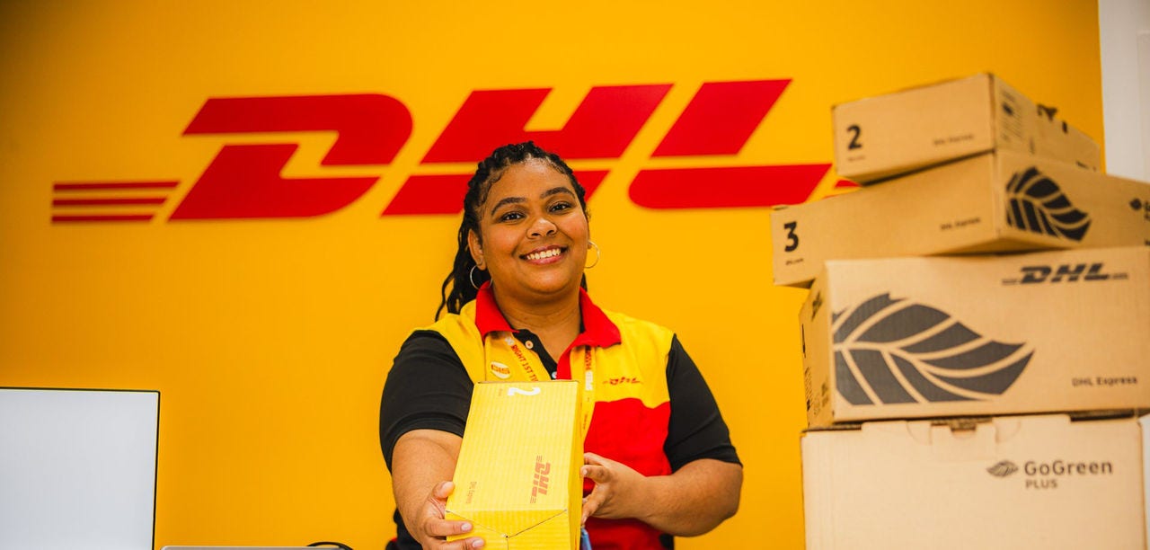 Atendente sorrindo no balcão de uma Loja DHL Express, entregando uma caixa DHL