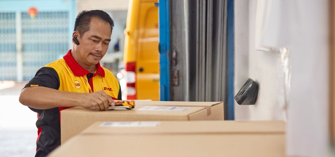 A DHL employee is in the warehouse of Thumbinthai, scanning the shipping labels of several brown parcels and preparing them for forwarding.