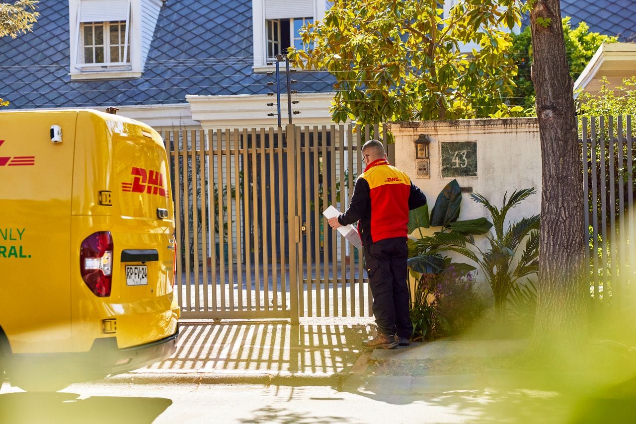 A male DHL employee is waiting at the closed gate of a private house after ringing the doorbell.