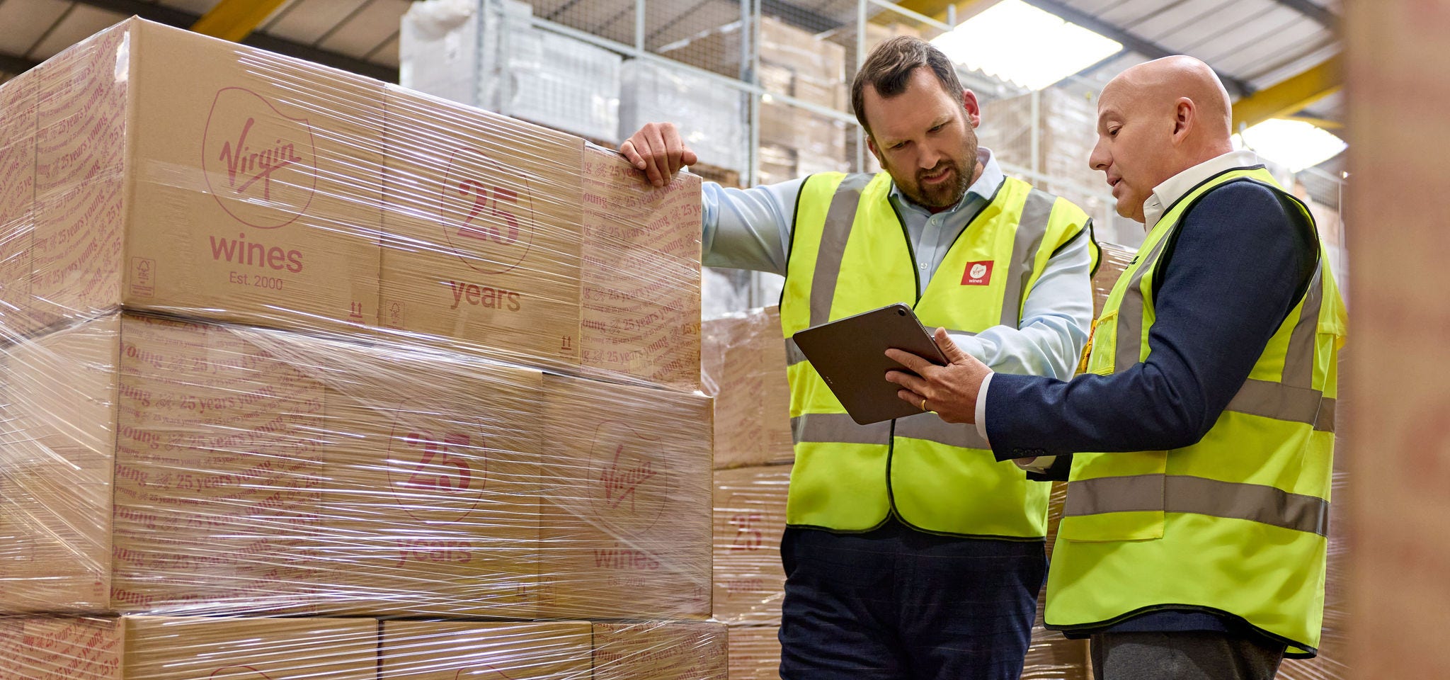 A male business customer is showing around a male DHL expert at a Virgin Wines warehouse, while they are talking about shipment options and looking together at a tablet.