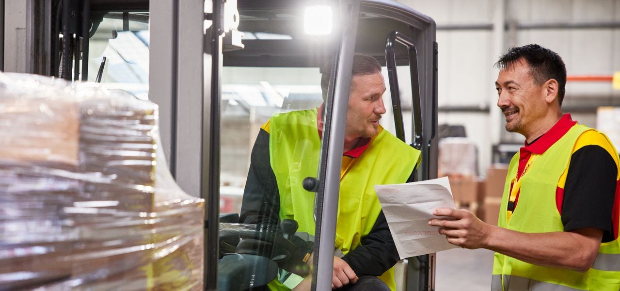 Employee in a fork lift talking to another employee who's holding papers.