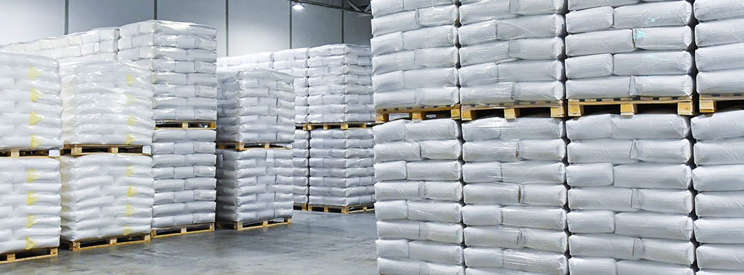 Large stacks of white bags, neatly stacked on wooden pallets and wrapped in transparent plastic. An industrial building or warehouse for storing food, raw materials, or other goods.