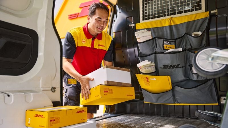 A male DHL employee loads several parcels into the loading zone of his electric DHL van