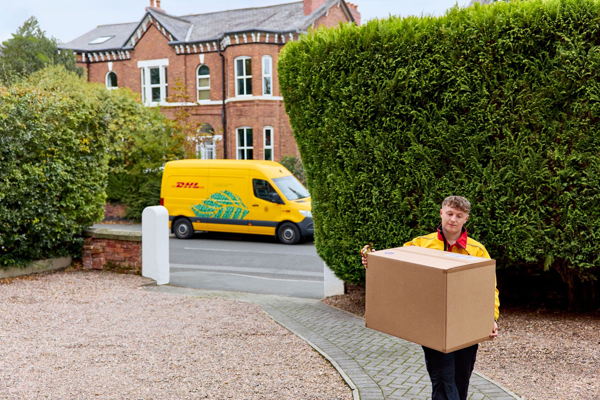 A male DHL employee with a large brown parcel walks through a front yard. A parked electric DHL van standing in the background.