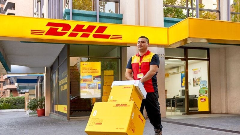 a male dhl employee walking outside a dhl service point with several parcels on a hand truck