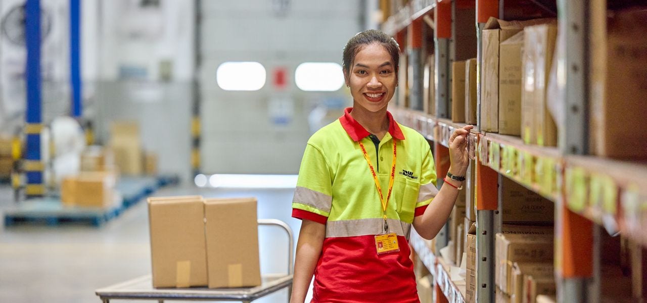 A female DHL employee stands next to a shelf in a multi-user warehouse.