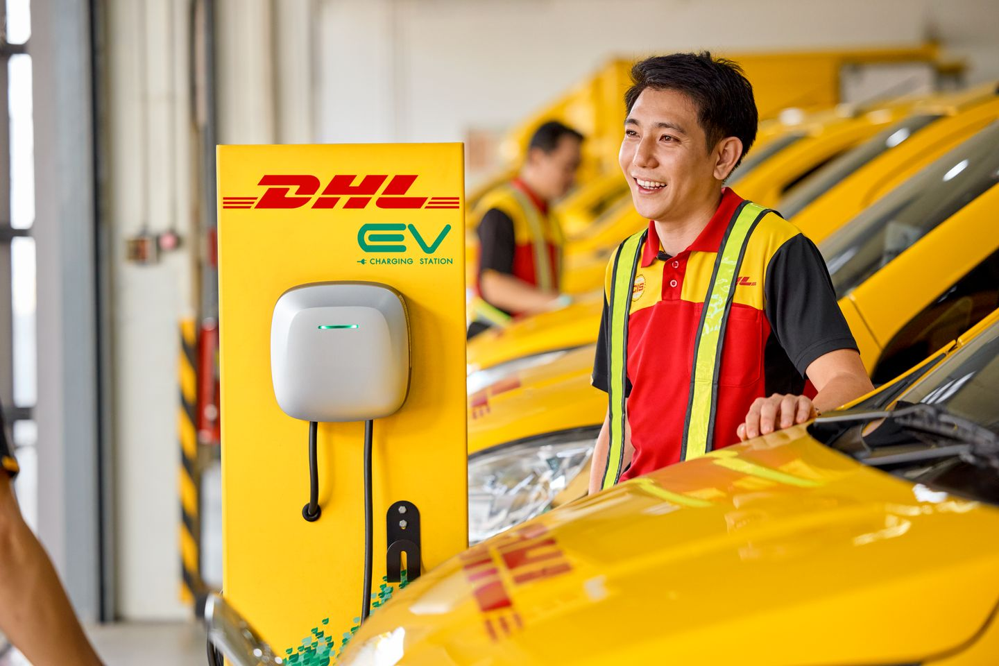 male dhl employee standing between electrical dhl van and dhl charging station