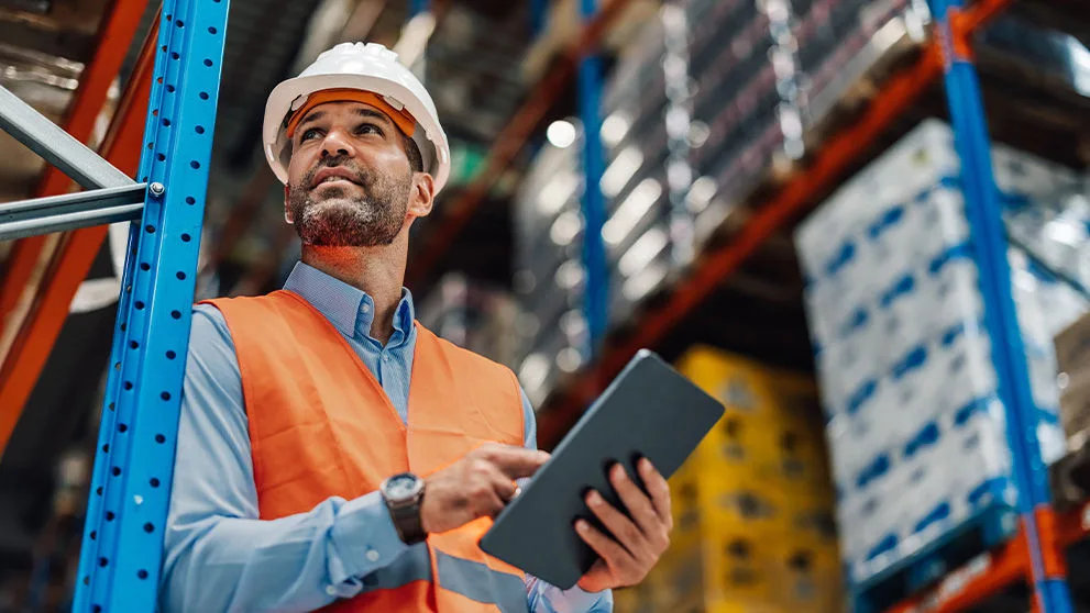 Warehouse manager wearing reflective vest and hardhat using digital tablet, controlling inventory and planning work in warehouse