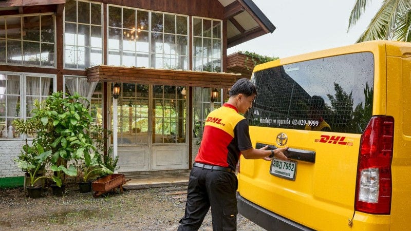 A DHL employee opens the back door of a DHL van in front of a remote house.