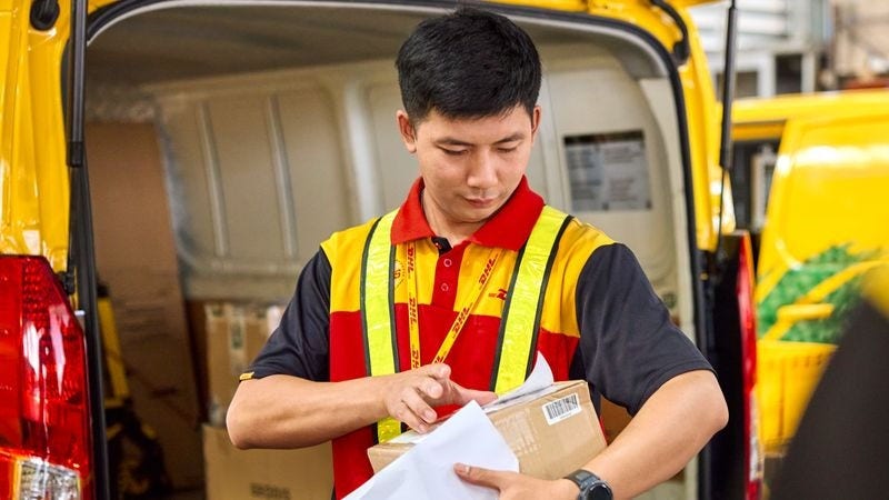 A DHL Express deliveryman attaches a label to a parcel in front of a van. 
