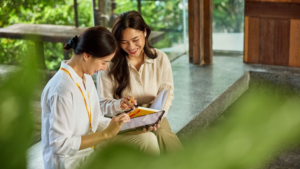 Two female DHL employees are sitting on a bench in a sustainable office, surrounded by plants, looking at a tablet together and chatting.