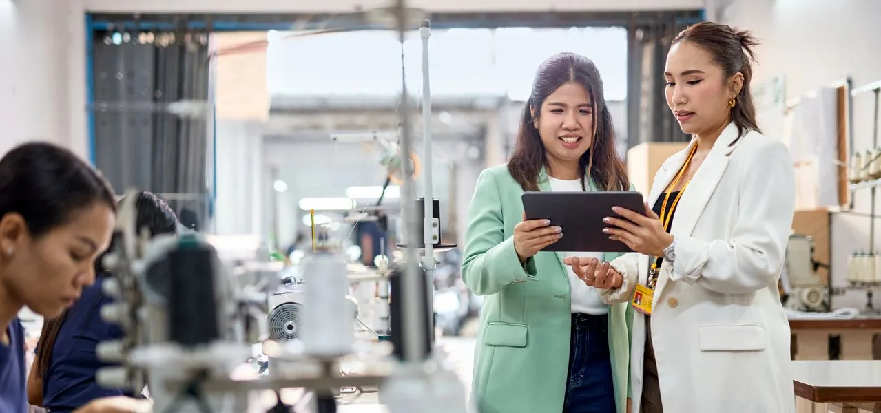 A female DHL expert is consulting a female business customer on site at Thumbinthai. Both are looking at a tablet, engaged in conversation. In the background, several seamstresses are working on sewing machines