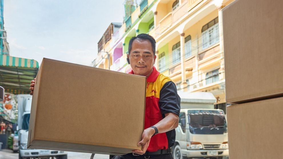 A male DHL employee loads a brown parcel into the cargo area of ​​his electric DHL delivery van.