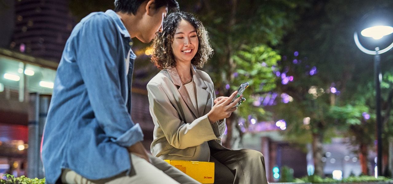 A female and a male customer with an express parcel are sitting on a bench in a city at night and looking at a smartphone.