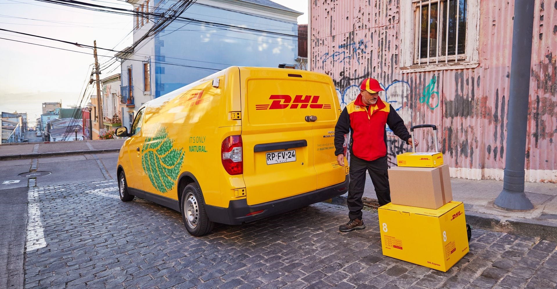 พนักงานชายของ DHL กําลังบรรทุกพัสดุบนรถบรรทุกมือข้างรถตู้ไฟฟ้า DHL ของเขา