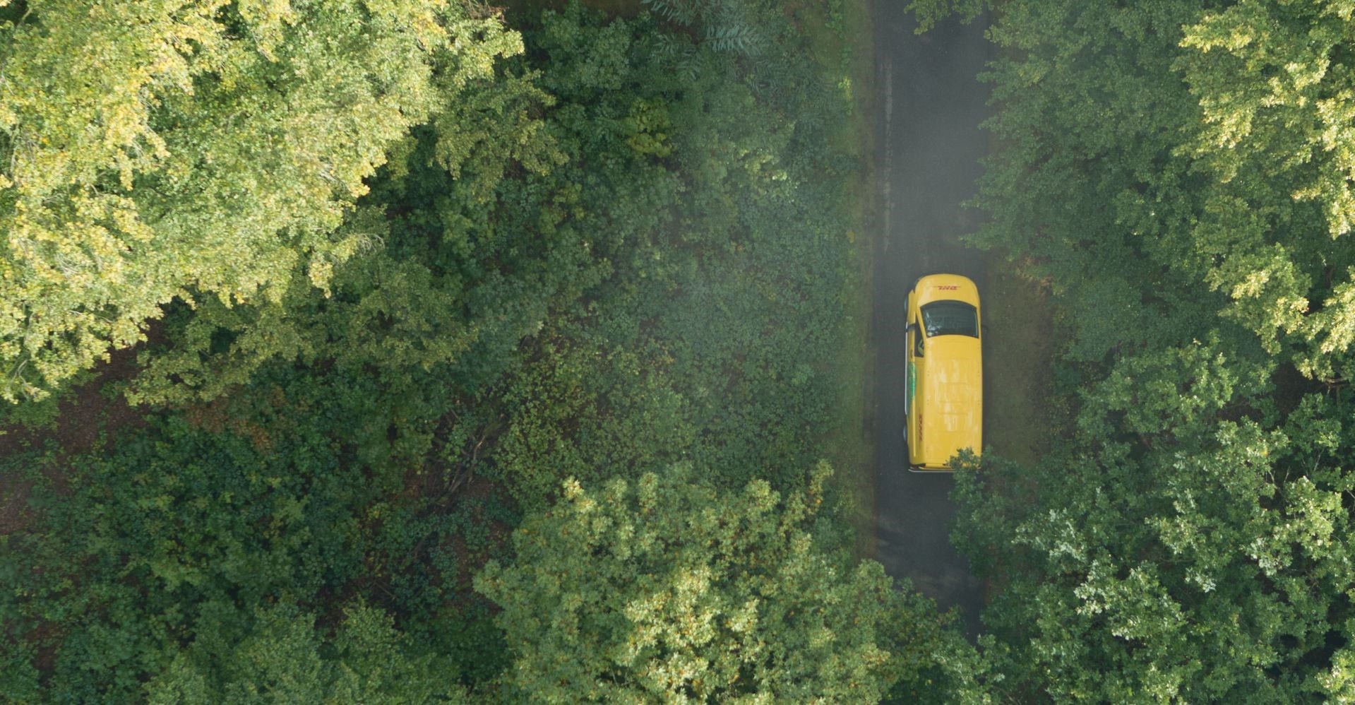 top down shot of a yellow dhl van driving on a road in the middle of a green forest