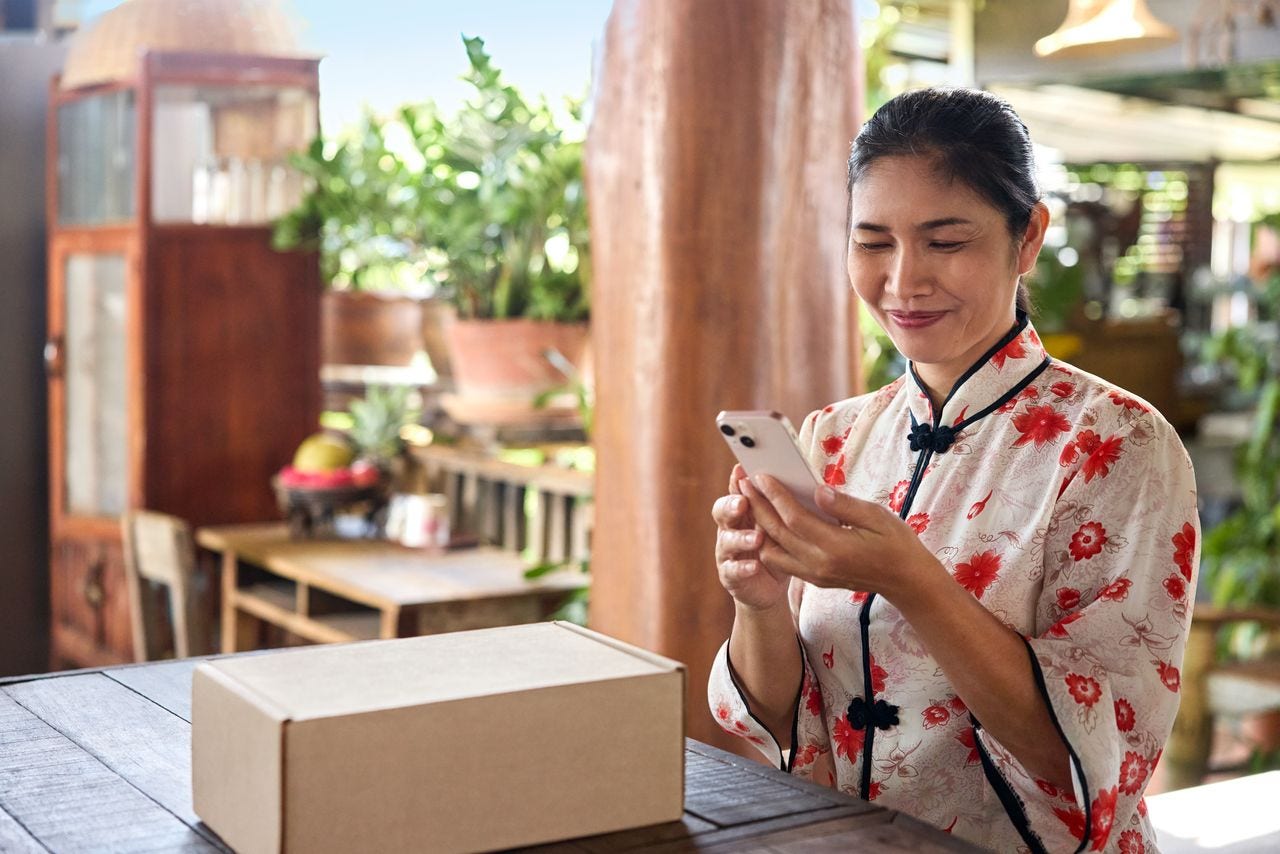 A female customer sits at the dining table in her traditional Thai home and is using her smartphone. Next to her on the table, a parcel is placed.