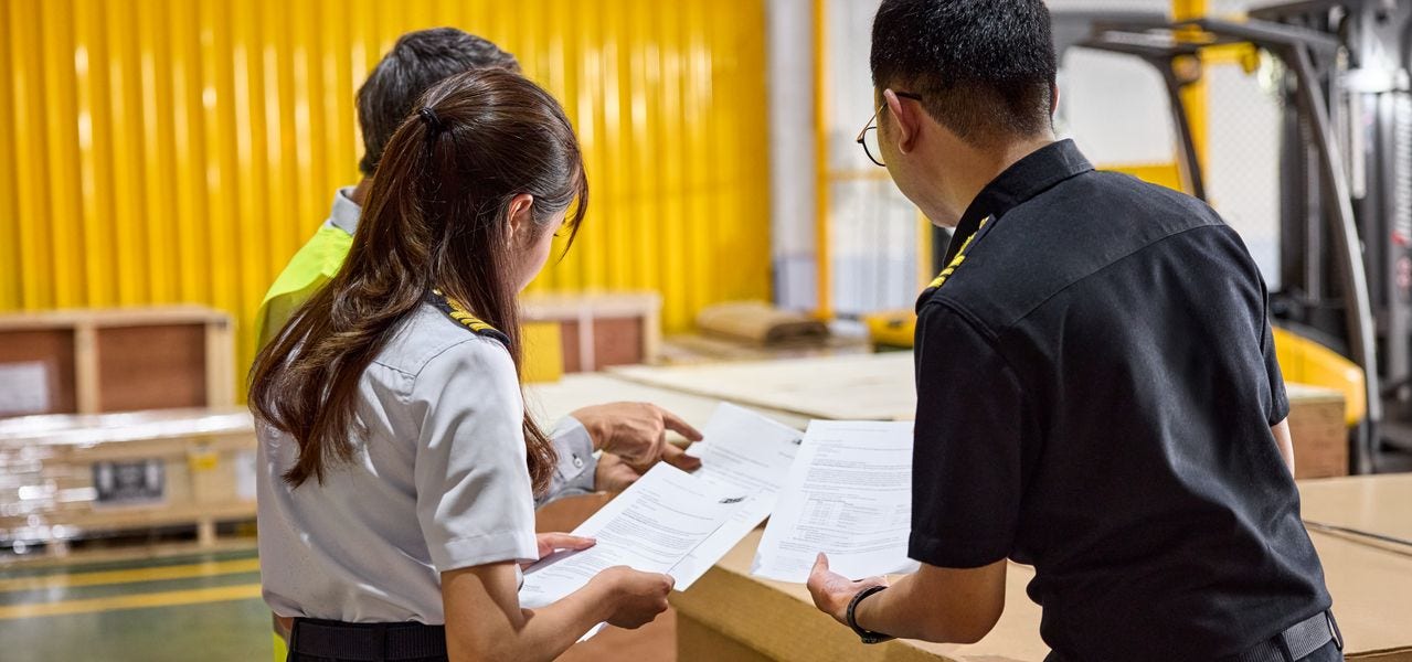 A male DHL expert inspects documents for a large transport box in a multimodal hub together with two customs officers.