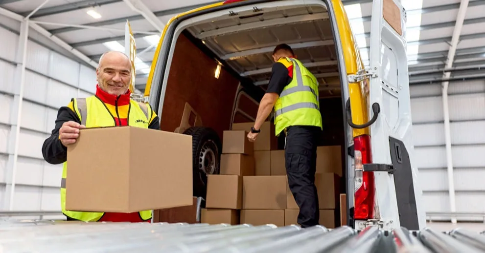 Two male DHL employees are unloading parcels from a DHL van onto a conveyor belt.