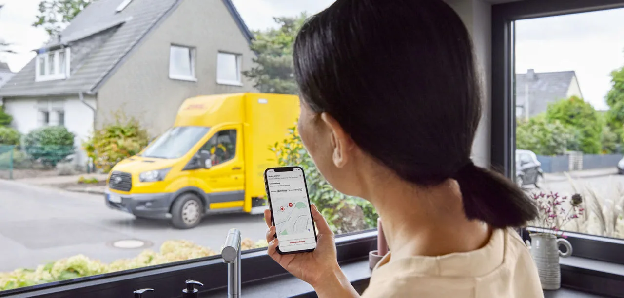 Woman looking out the window at a DHL truck while using the DHL app to track its arrival