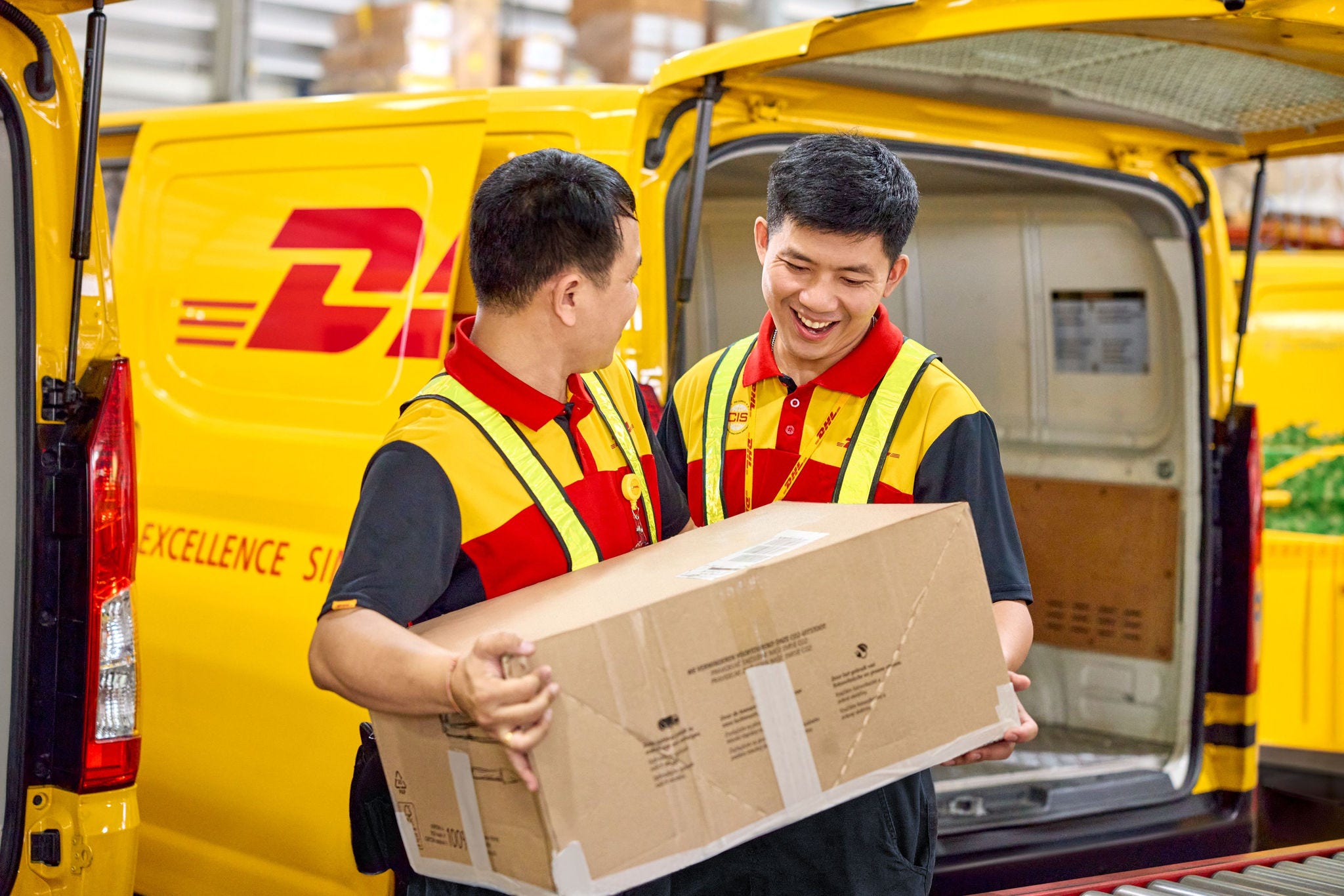 Two male DHL employees load a large parcel together into an electric DHL van. 