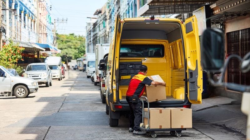 Seorang karyawan DHL laki-laki memuat paket cokelat ke area kargo van pengiriman DHL listriknya.