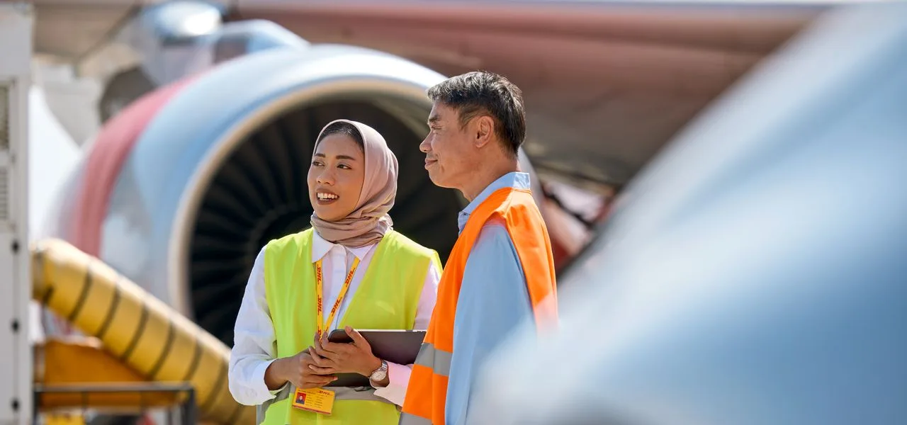 A female DHL expert and a male DHL customer having a conversation on the airfeld in front of a plane which is loaded with cargo