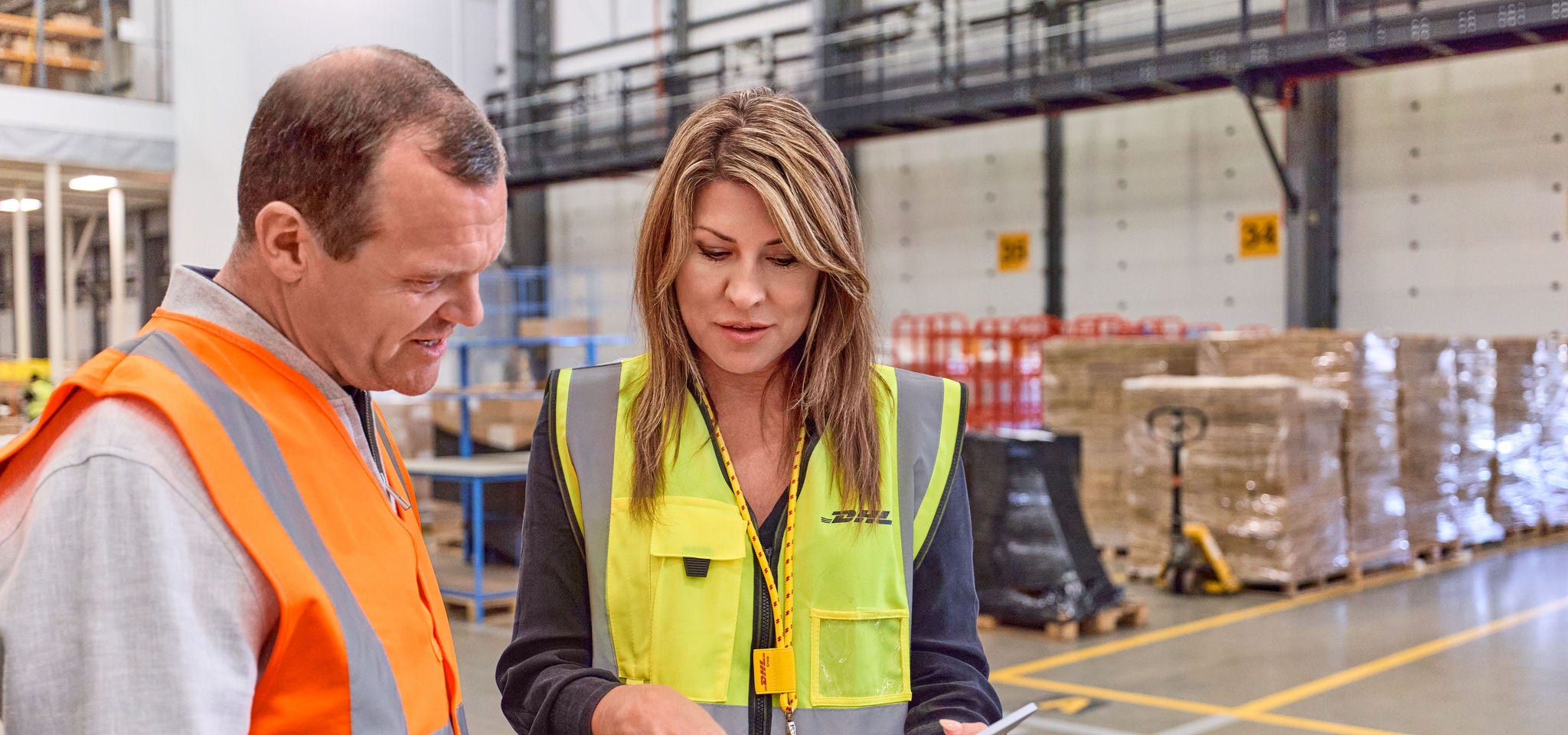 A female DHL expert is consulting a male business customer at a warehouse. Both are looking at a tablet, engaged in conversation.
