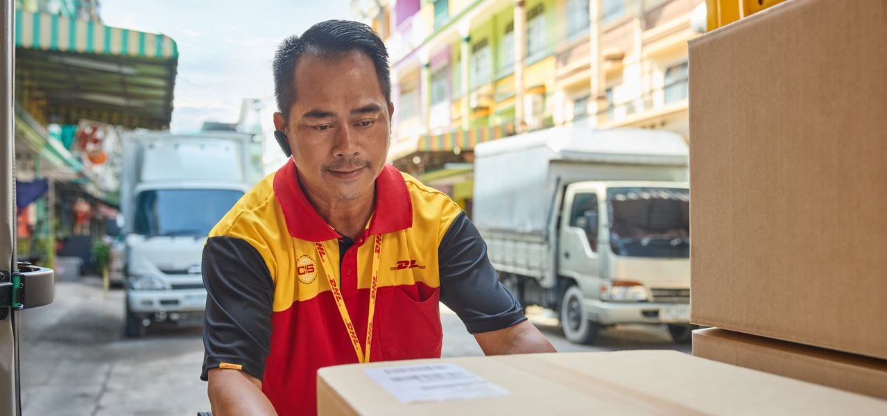 A male DHL employee loads a brown parcel into the cargo area of ​​his electric DHL delivery van. In the background, a narrow street lined with residential buildings and small shops can be seen.